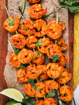 A pile of grilled spicy shrimp skewers topped with chopped herbs with lime segments nearby.