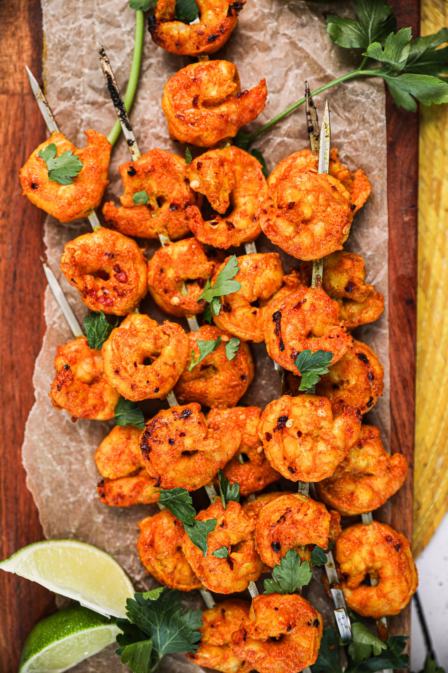 A pile of grilled spicy shrimp skewers topped with chopped herbs with lime segments nearby.