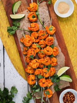 Birdseye view of pile of grilled spicy shrimp skewers topped with chopped herbs with lime segments, sprigs of herbs and ramekins of garlic powder and chilli flakes nearby.