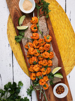 A pile of grilled spicy shrimp skewers topped with chopped herbs with lime segments, sprigs of herbs and ramekins of garlic powder and chilli flakes nearby.