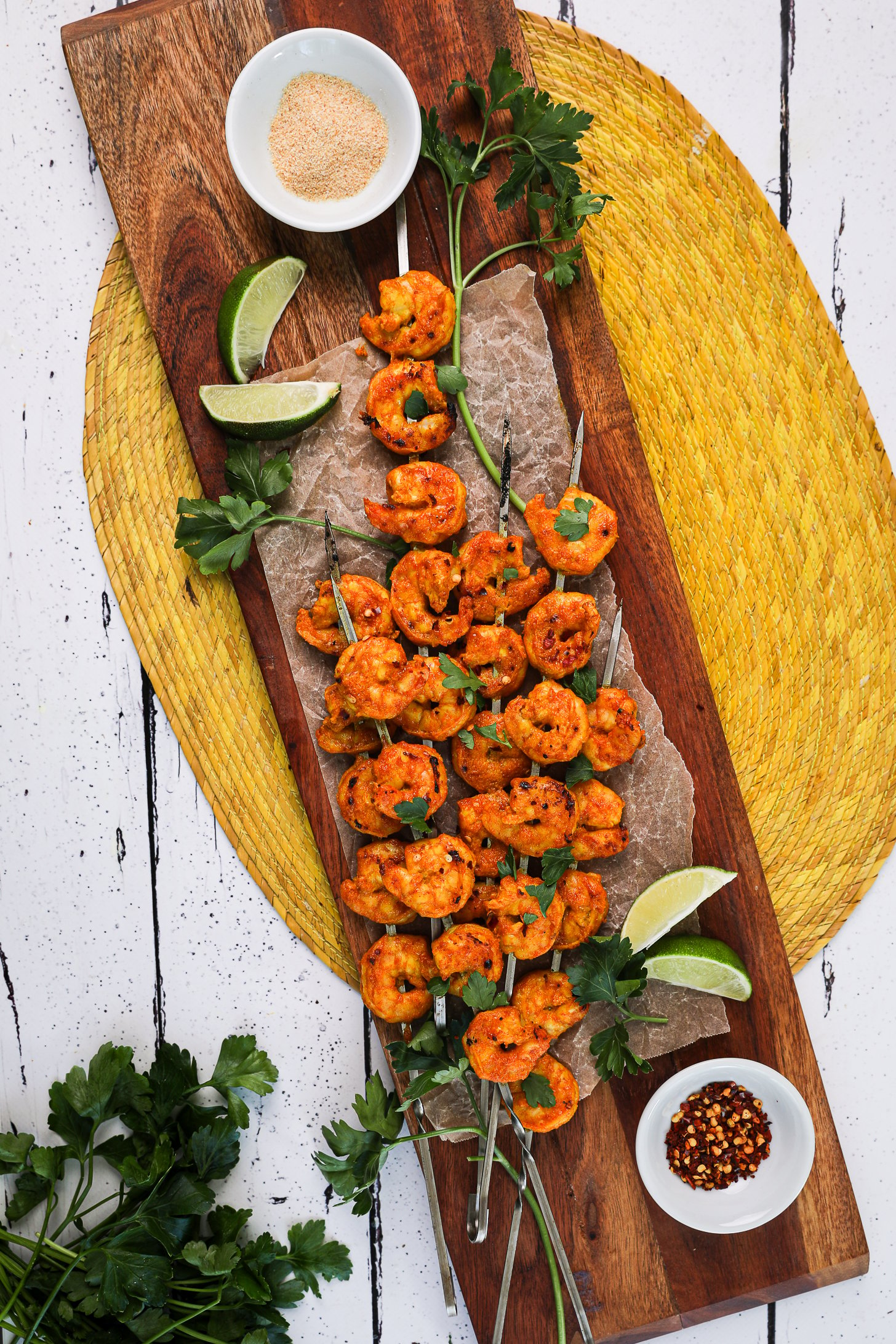 A pile of grilled spicy shrimp skewers topped with chopped herbs with lime segments, sprigs of herbs and ramekins of garlic powder and chilli flakes nearby.