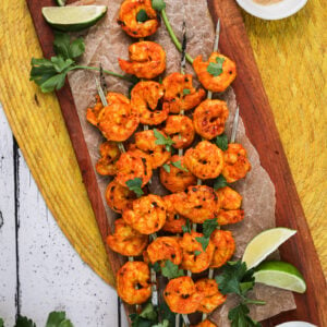 Birdseye view of pile of grilled spicy shrimp skewers topped with chopped herbs with lime segments, sprigs of herbs and ramekins of garlic powder and chilli flakes nearby.